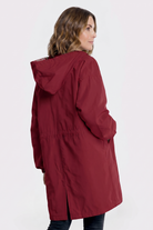 A person with shoulder-length wavy hair is seen from behind wearing the Icon Windbreaker Rain Jacket, a long, hooded red coat with a bottom slit and loose fit, standing against a plain light background.