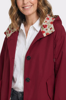 A woman smiles softly, wearing the Icon Windbreaker Rain Jacket in maroon with black buttons over a white shirt. The hood is lined with red floral fabric, and her wavy, shoulder-length hair frames her face.