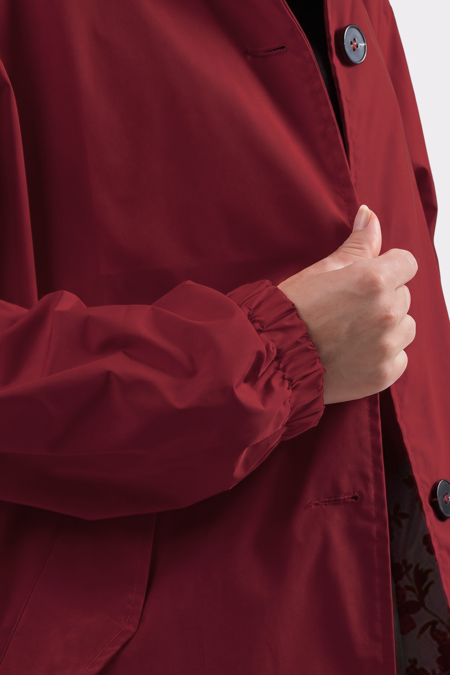 A person wearing the Icon Windbreaker Rain Jacket in red is shown from the shoulders down, holding the edge of the jacket with their right hand. The jacket features large black buttons and an elasticated sleeve.