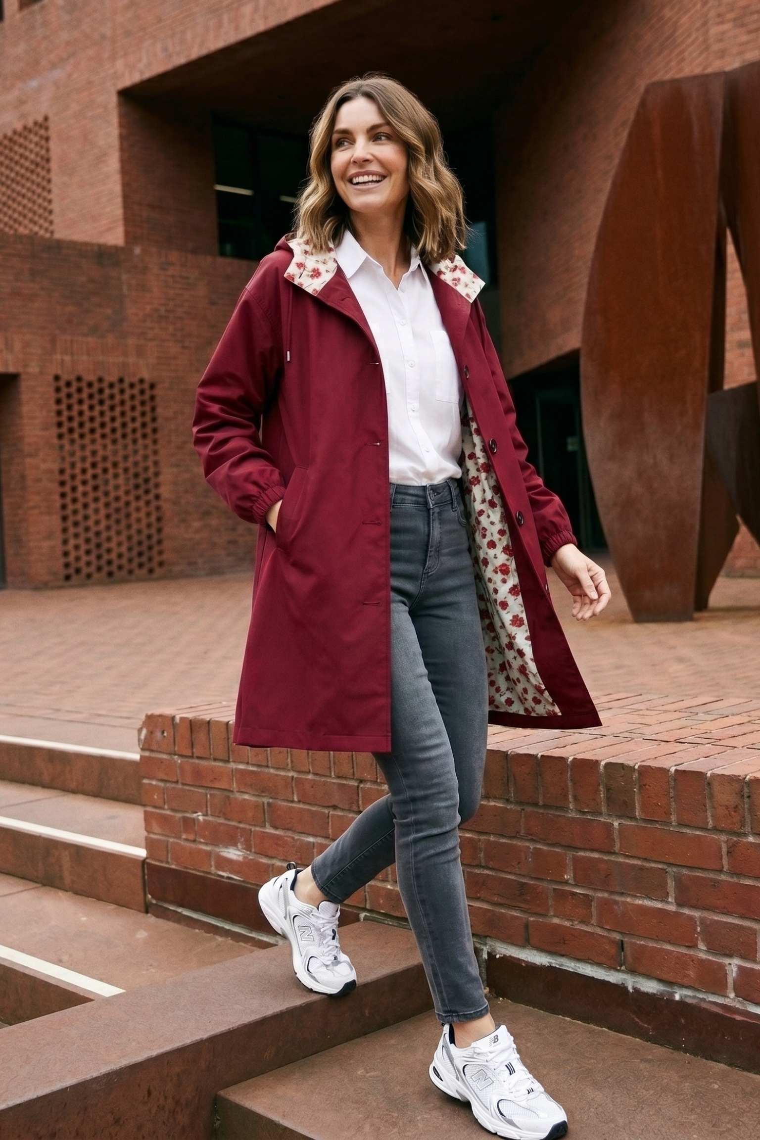 A woman with shoulder-length hair and a smile walks down outdoor steps in the Icon Windbreaker Rain Jacket, a white shirt, gray jeans, and white sneakers. Red brick buildings are visible in the background.