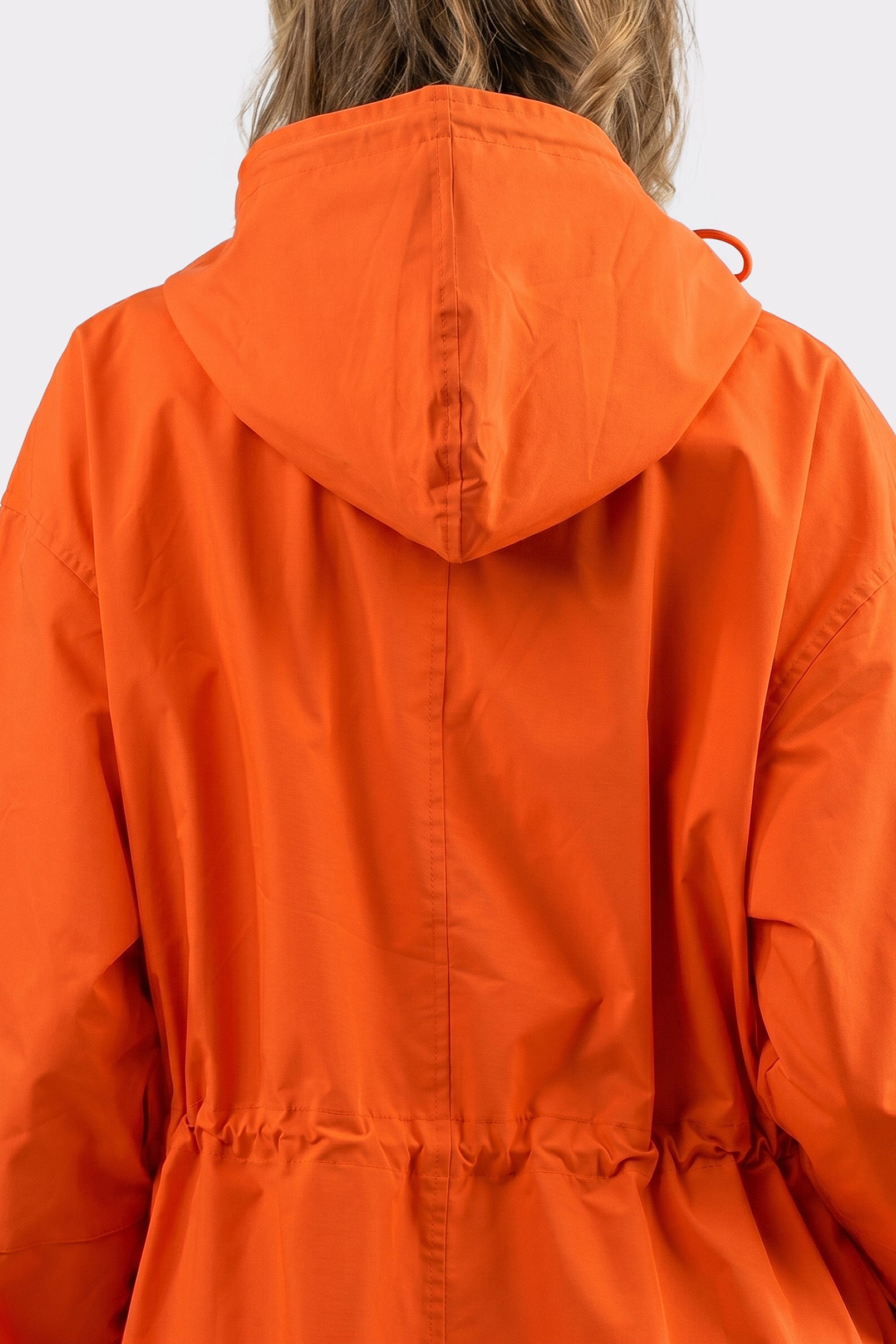 A person with wavy, light brown hair is seen from behind wearing the Icon Windbreaker Rain Jacket in orange, featuring a cinched waist. The background is plain and light-colored.