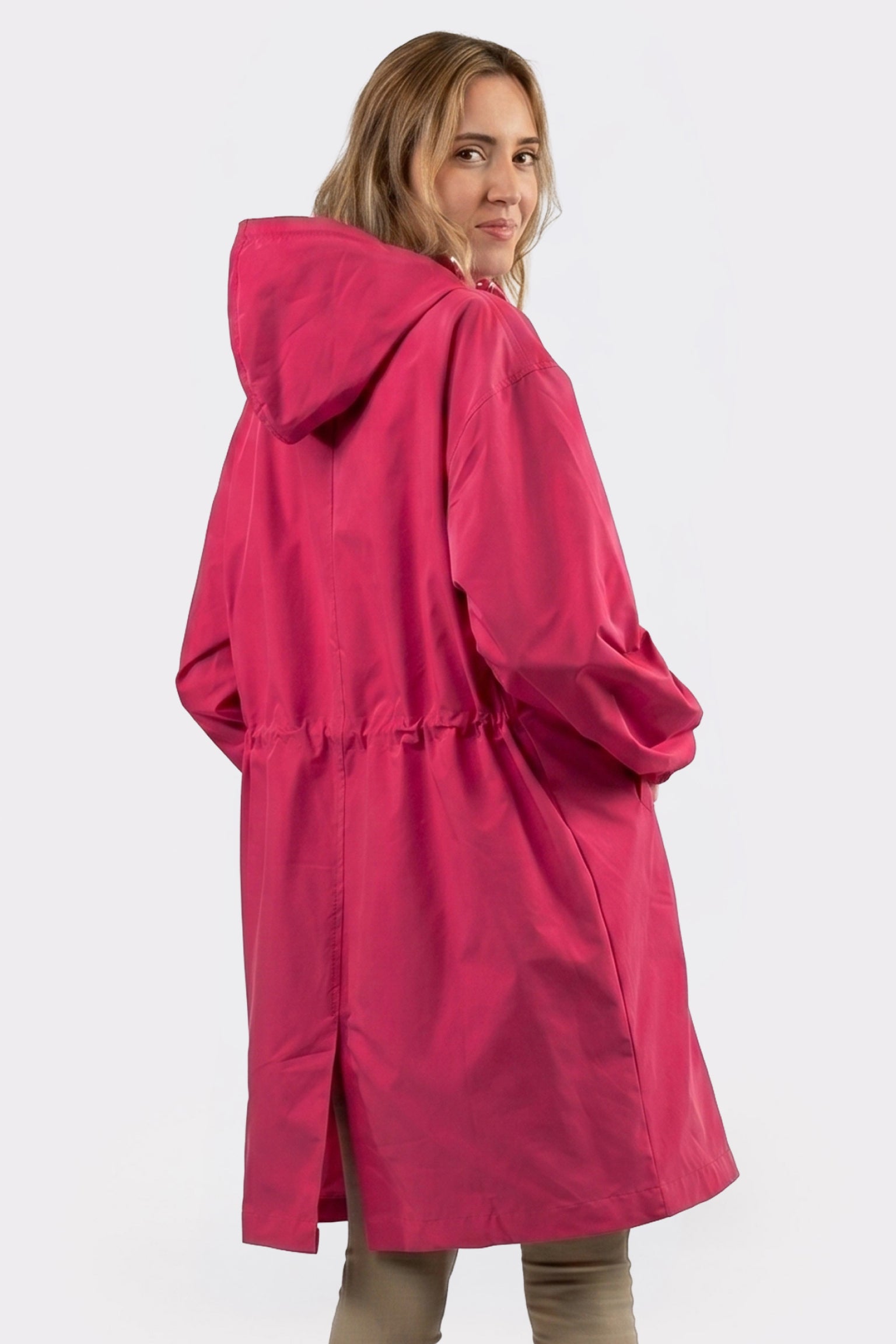 A woman with loose light brown hair stands slightly turned away, looking over her shoulder and wearing the Icon Windbreaker Rain Jacket in bright pink with her hands in the pockets, set against a plain white background.