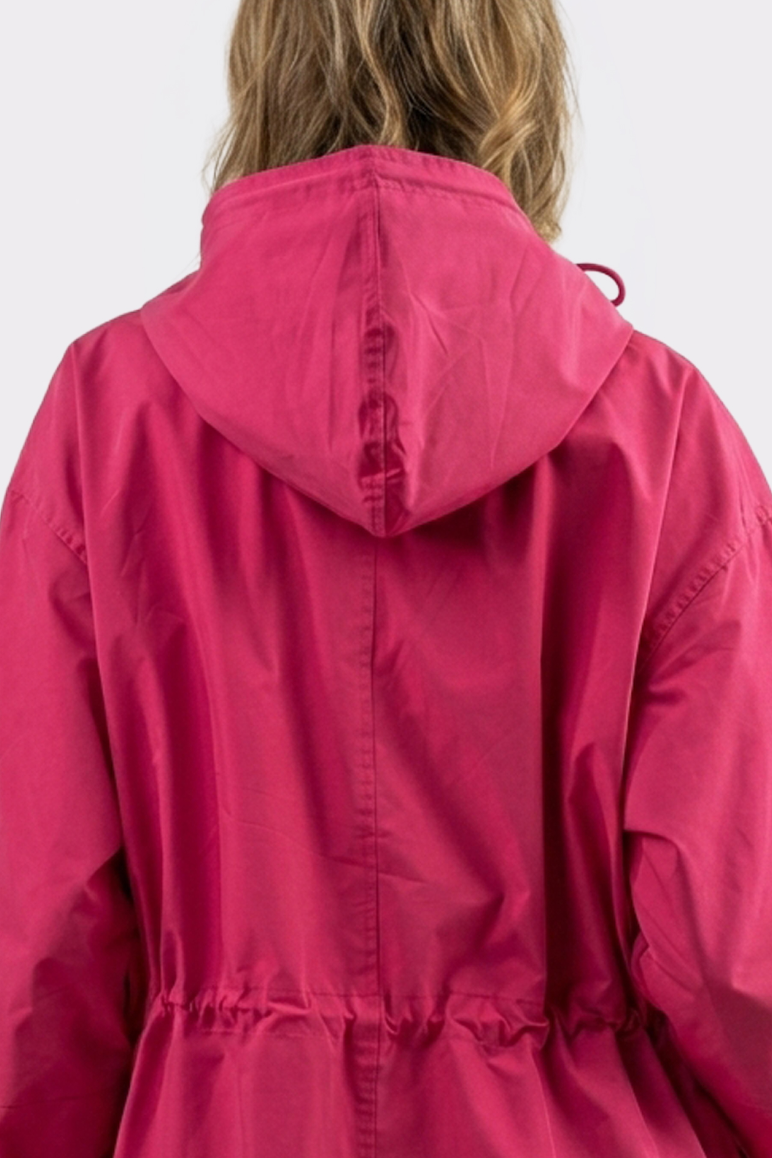 A person with light brown hair seen from behind wears the Icon Windbreaker Rain Jacket—a bright pink hooded style with a waist drawstring. The hood is up (not covering the head) and the background is plain light gray.