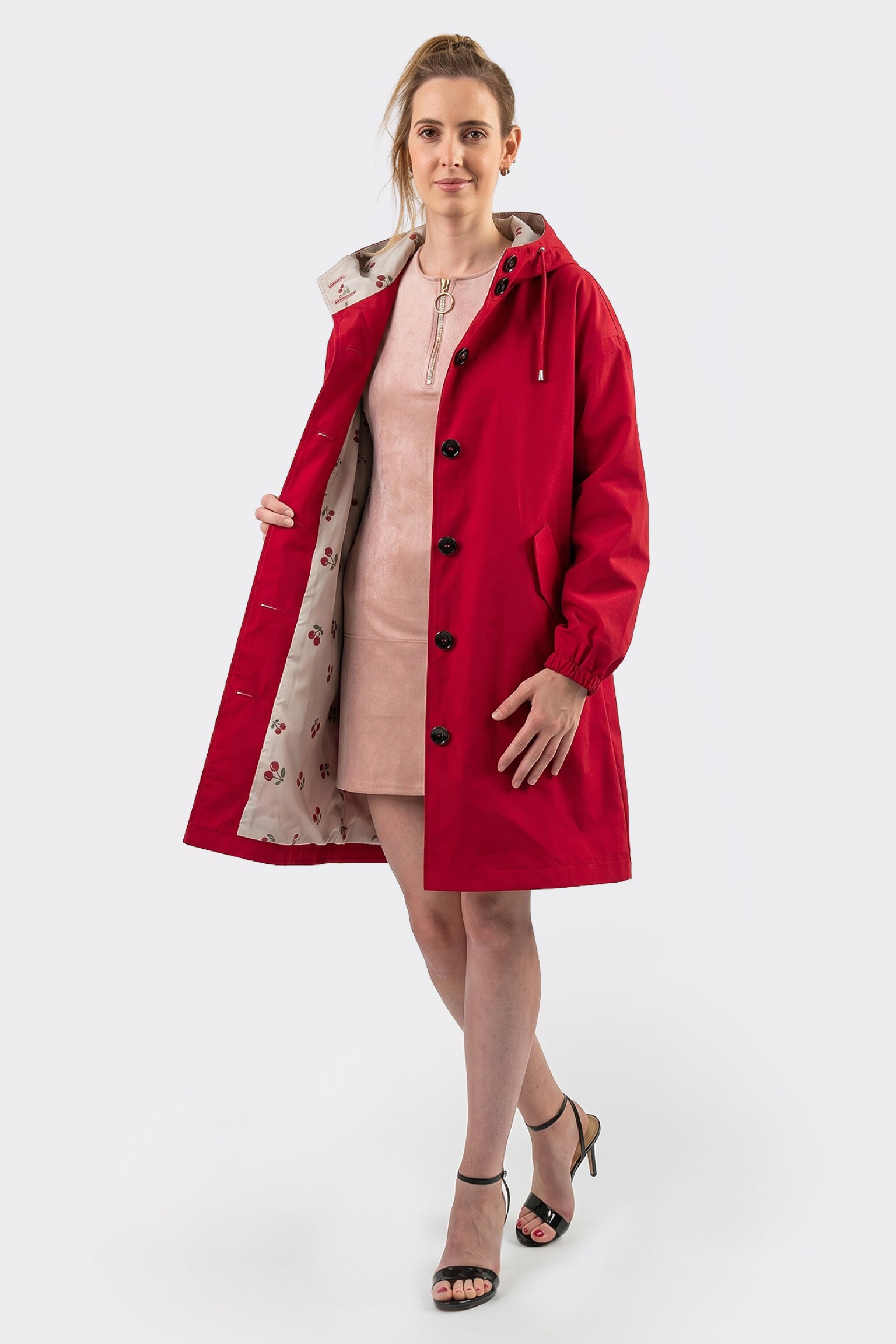 A woman stands against a white background wearing a light pink dress and the Icon Windbreaker Rain Jacket, styled open to show its decorative lining. She pairs this with black high-heeled sandals and faces forward, one hand holding the jacket open.