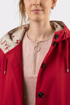 A woman wears the Icon Windbreaker Rain Jacket with a red hood, cherry pattern lining, and a light pink zip-up top underneath. The image is cropped from her shoulders to chest.