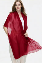 A woman with long light brown hair stands against a plain background, wearing the looselyboho Velvet Merlot 100% Cashmere Scarf over a white top and light pants, looking at the camera with a slight smile.