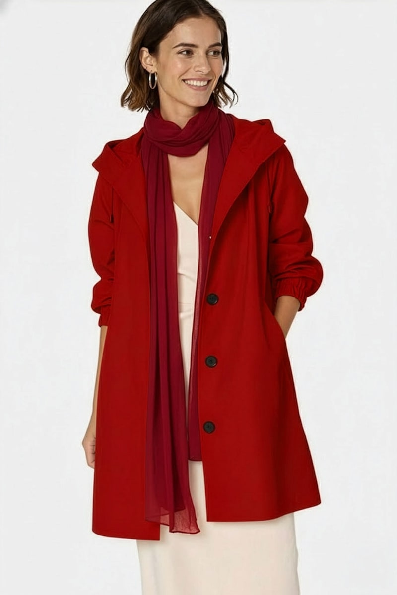A woman in a long red coat over a light dress wears the looselyboho Velvet Merlot 100% Cashmere Scarf, standing with her hands in her coat pockets and gazing slightly to her left.