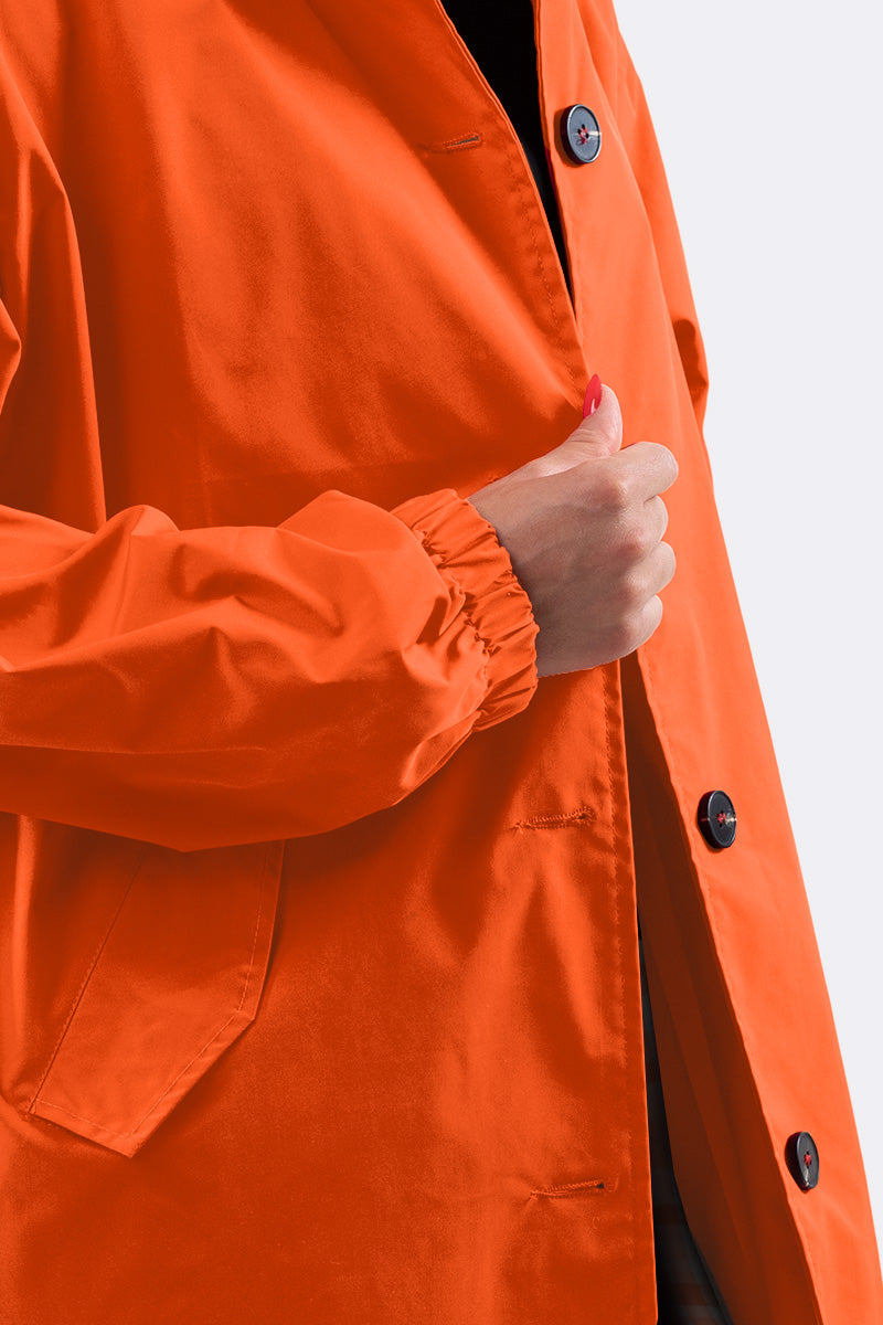 A person wearing a PUMPKIN ORANGE WINDBREAKER by looselyboho, featuring black buttons. Their hand is partially tucked into the lightweight jacket, revealing a sleeve with an elastic cuff against a plain, light-colored background.