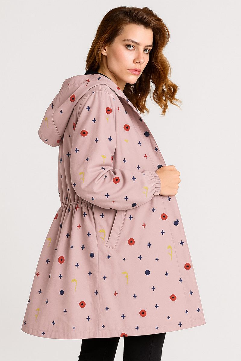 A woman stands in profile with one hand in her pocket, wearing the looselyboho DOT & BLOOM WINDBREAKER—a light pink windbreaker with multicolored dots and crosses—against a plain, light background, looking toward the camera.