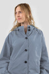 A person with long blonde hair is wearing "The Windbreaker Jacket" in powder blue. The jacket, named POWDER BLUE WINDBREAKER, features large black buttons and a high collar. The individual is looking slightly to the side, set against a plain and light-colored background.