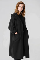 A woman with shoulder-length light brown hair stands against a plain background, wearing the looselyboho Black One Size Fits All Oversized Water Resistant Windbreaker Jacket. With her hands in the pockets, she gazes slightly to the side with a neutral expression.