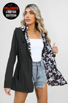 A woman with long, wavy blonde hair is wearing a black FLORALSHIFT COAT: THE REVERSIBLE RAINCOAT by looselyboho, featuring a floral-patterned lining. Underneath, she sports a white tube top and high-waisted denim shorts. She holds the coat open to showcase its deep pockets and intricate lining. On the top left corner of the coat is a sticker that reads "Pre-Order Limited Edition.