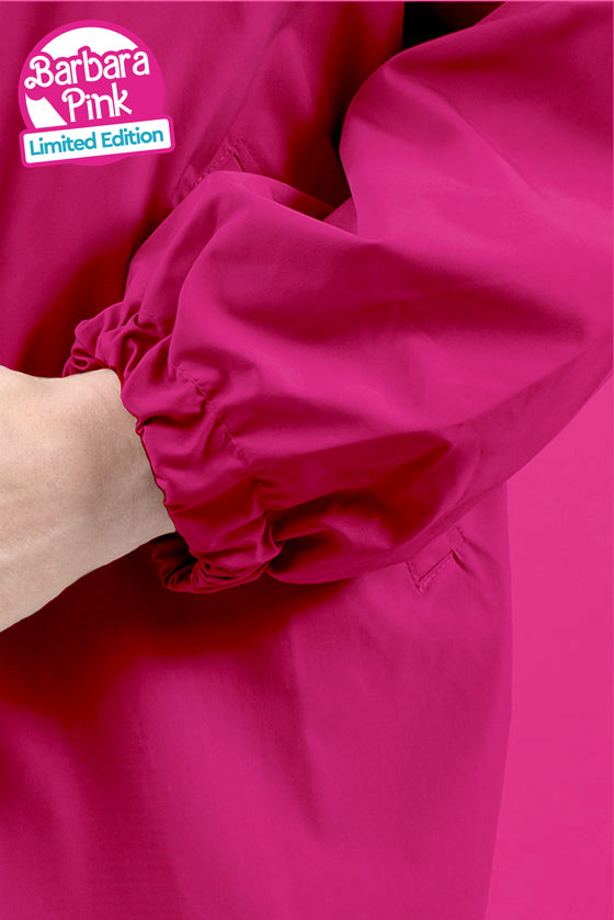 Close-up of an arm wearing a bright pink BARBARA PINK WINDBREAKER from looselyboho with an elasticated cuff. The lightweight, water-resistant fabric appears glossy. In the upper left corner, there is a logo with the words "Barbara Pink" and "Limited Edition." The background matches the jacket's vibrant shade of pink.