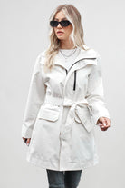 A woman with long, blonde hair is wearing a BELTED FROST WHITE WINDBREAKER by looselyboho, black sunglasses, and dark jeans. She stands against a plain gray background, with one hand holding the front of her coat. She wears a white top underneath and layered necklaces.