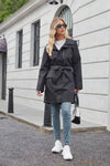 A woman with long blonde hair wears a looselyboho BELTED MIDNIGHT BLACK WINDBREAKER tied at the waist, light blue jeans, and white sneakers. She carries a small black handbag and sports dark sunglasses. She is walking down a grey street with a white building in the background.