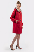 A person stands against a plain light background wearing the Icon Windbreaker Rain Jacket, a knee-length red hooded coat with black buttons and side pockets, paired with black high-heeled sandals.