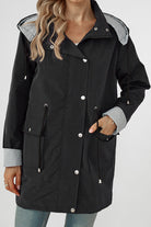 A person with wavy blonde hair is wearing a black waterproof raincoat jacket with silver snap buttons and drawstrings. The looselyboho STRIPED CARBON BLACK WINDBREAKER JACKET, featuring a striped lining at the cuffs and hood, has a lightweight design. The person has one hand in their pocket and is dressed in light blue jeans.