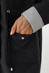 A person's hand is partially in a front pocket of a dark, lightweight **looselyboho STRIPED CARBON BLACK WINDBREAKER JACKET**. The sleeve of the jacket is rolled up, revealing a striped inner lining. A black drawstring with a silver metal tip hangs next to the pocket. The fingernails are neatly manicured and painted light pink.