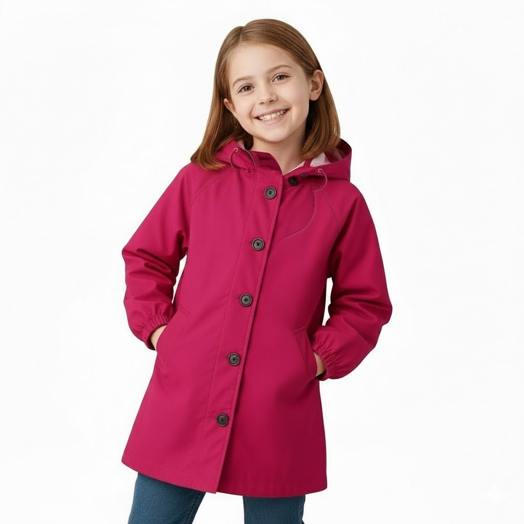A young girl with straight light brown hair smiles at the camera, hands in the pockets of a looselyboho Bright Berry Kids’ Windbreaker, paired with blue pants against a plain white background.
