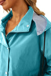 A close-up of a person wearing a fashionable looselyboho STRIPED TURQUOISE OASIS WINDBREAKER JACKET with a zippered and buttoned front. The collar of the lightweight jacket is partially open, revealing a lining with blue and white stripes. The person has light-colored hair and is shown from the shoulders up.