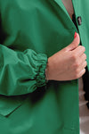 A person wearing an EMERALD GREEN WINDBREAKER by The Windbreaker Jacket is shown from the shoulder to the waist. Their hand is positioned near the jacket's black button, gripping the lapel. The sleeve is gathered at the wrist. The person has a neutral-colored garment underneath and is wearing a bracelet.